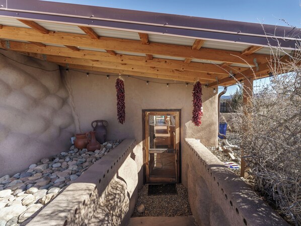 A rustic outdoor area with a wooden pergola, stone wall, and a door leading inside. There are two clay pots and a hanging red decoration.