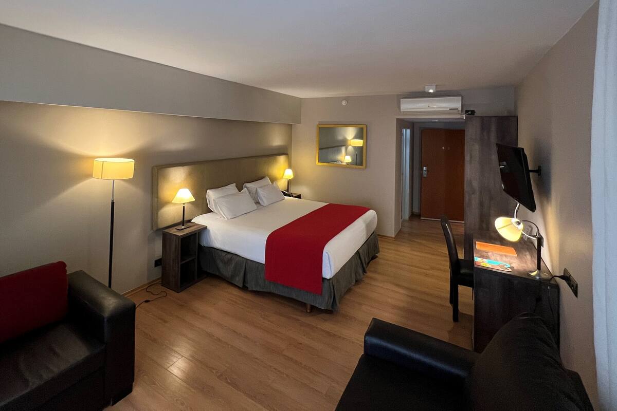 A hotel room with a bed, bedside lamps, a desk with a chair, a red throw, and a mirror on the wall.