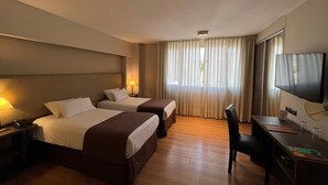 Ein Hotelzimmer mit zwei Betten, einem Schreibtisch, einem Stuhl, einem Fernseher und einem Fenster mit Vorhängen.