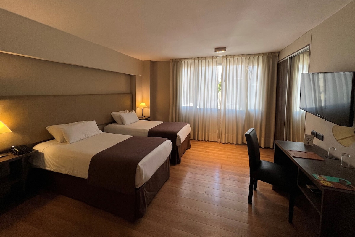 A hotel room with two beds, a desk, a chair, a TV, and a window with curtains.