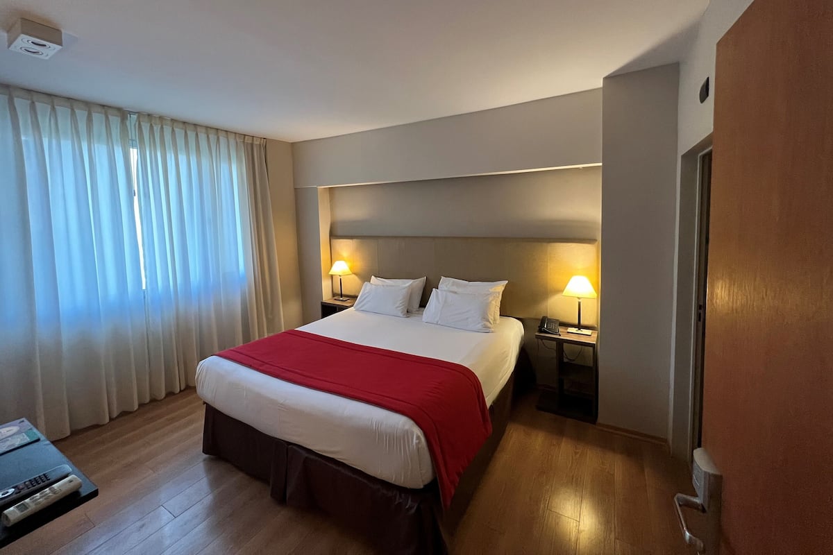A hotel room with a large bed, two bedside lamps, a red throw blanket, a wooden floor, and a window with curtains.