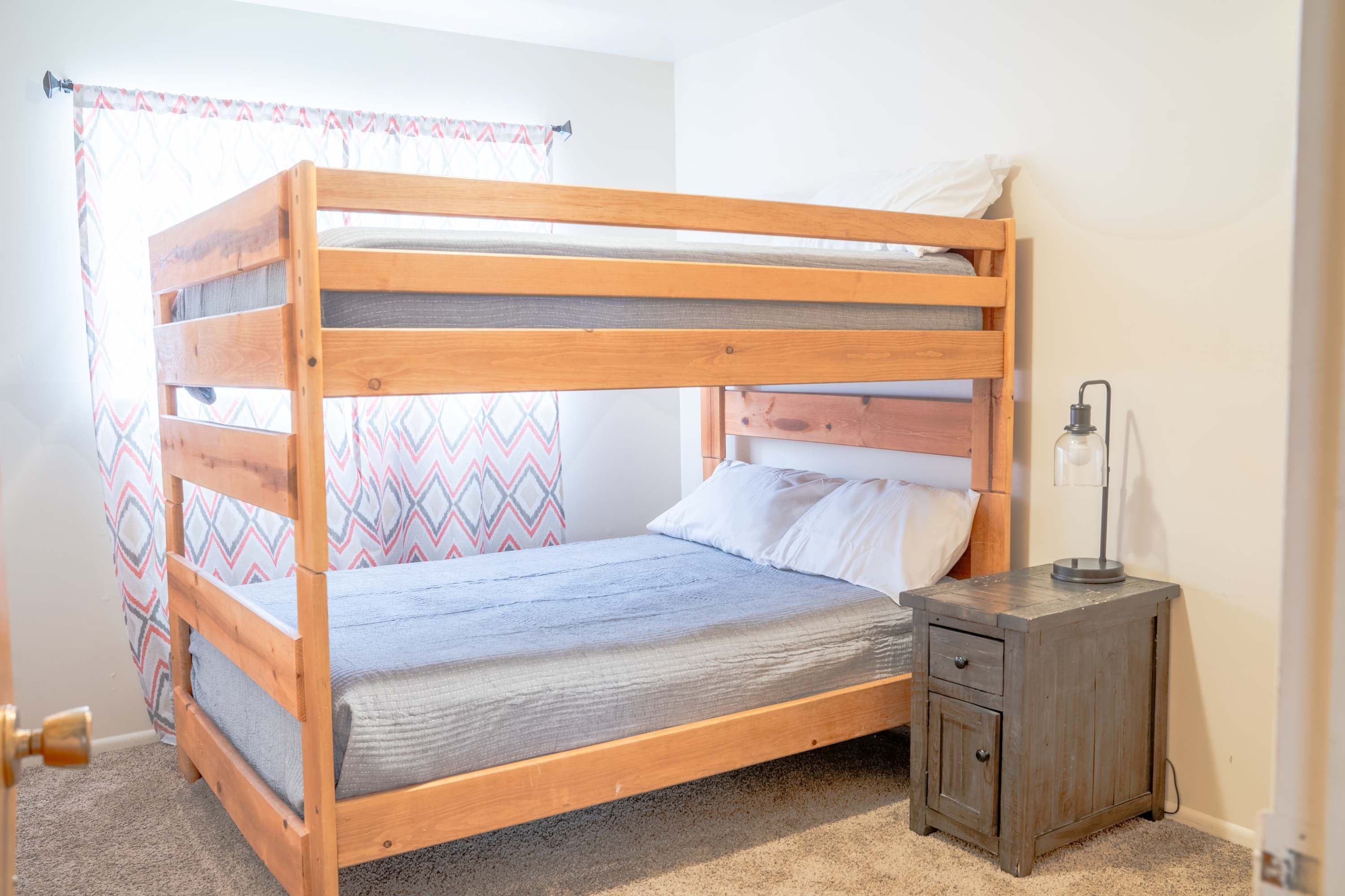 A bunk bed with a wooden frame, a bedside table with a lamp, and a patterned curtain.