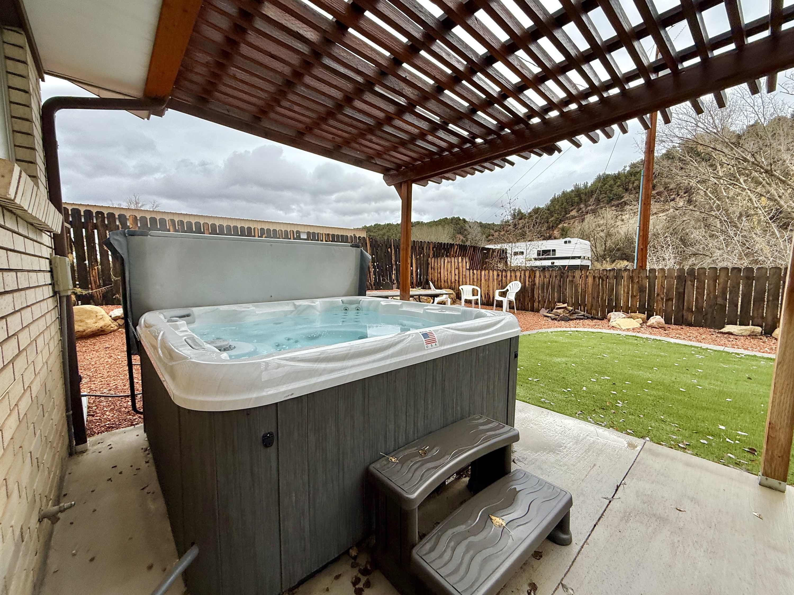 A hot tub under a wooden pergola in a backyard with a wooden fence, a grassy lawn, and a tree.