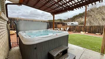 A hot tub under a wooden pergola in a backyard with a wooden fence, a grassy lawn, and a tree.