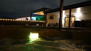 A hot tub area with a wooden deck, a covered seating area, and a view of a house with illuminated windows. - Spacious 4-bedroom house in Miami with Hot Tub, Lake, Kayaks, and Pool table (Miami)