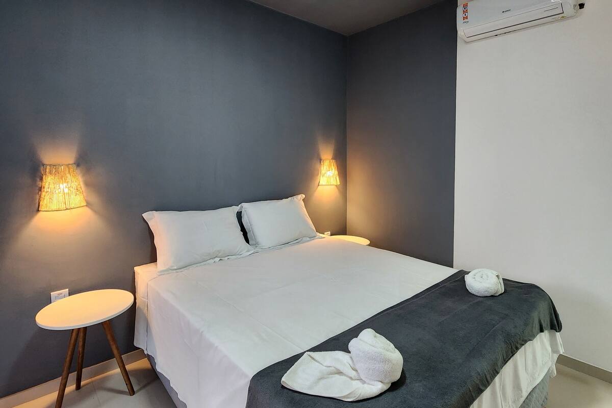 Una cama con ropa de cama blanca, un edredón gris oscuro y dos toallas sobre ella. A cada lado de la cama hay dos lámparas fijadas a la pared, y una mesita redonda con una lámpara al lado.
