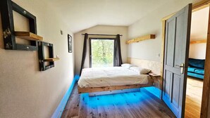A narrow room with a bed, wooden beams, a window with curtains, and a door leading to another room.