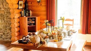 A wooden cabin interior with a dining table set for breakfast, a stone wall, and a window with curtains.