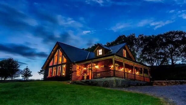 A large log cabin with a porch and multiple windows, surrounded by a grassy area and trees.