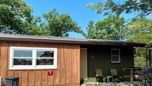 A small cabin with a wooden exterior, a window, and a door. There are two chairs outside and a table with a grill. - Lakeside Condo | Calm Waters Resort | Boat Slips & Rentals | FREE Kayaks & Paddleboards | Guided Fishing & Lake Tours! (Branson)