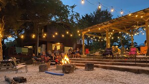 An outdoor seating area with a fire pit, surrounded by chairs and tables, under a wooden pergola with string lights.