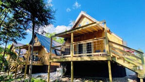 A wooden cabin with a balcony and a metal roof, surrounded by trees and gravel.