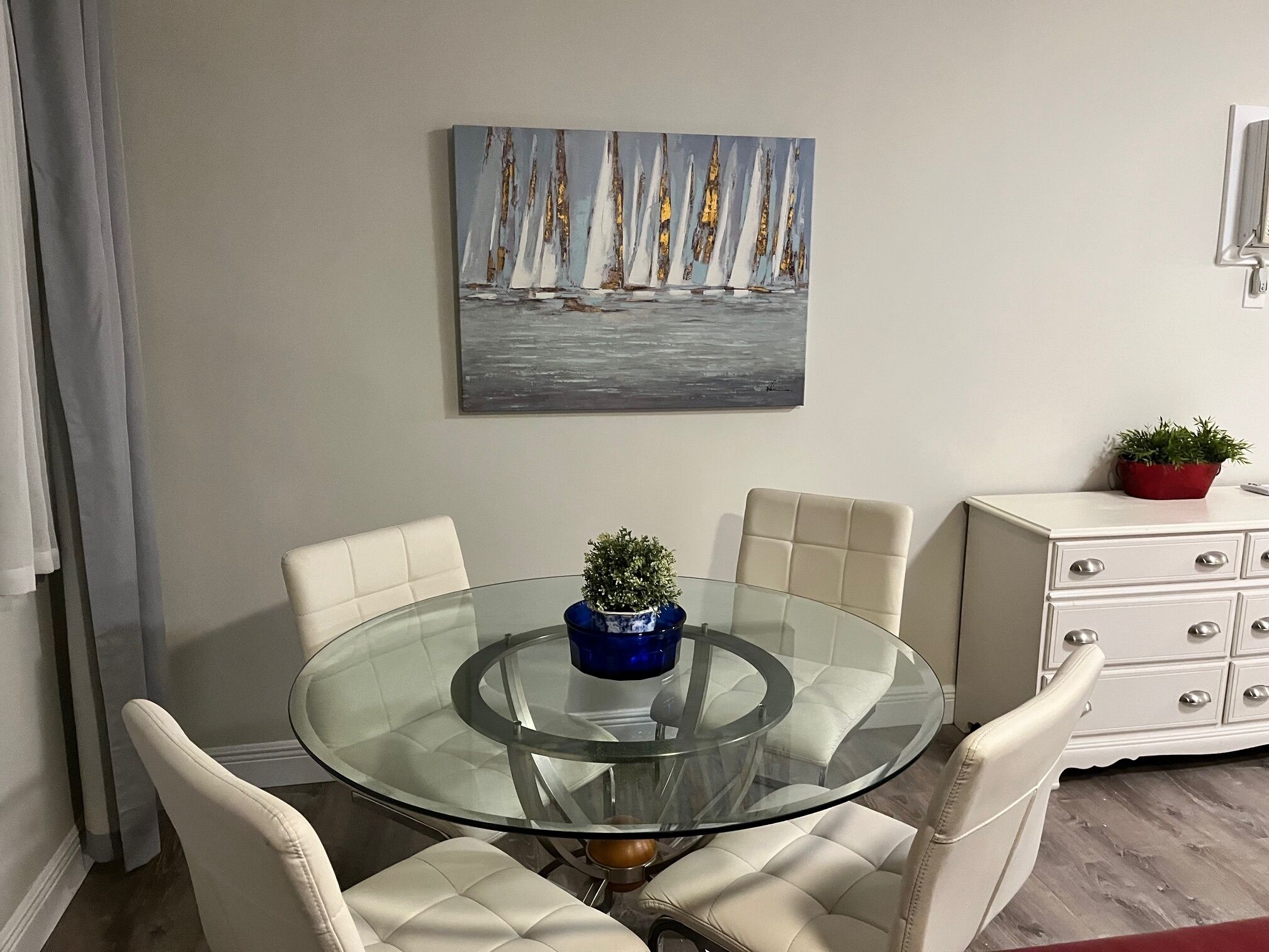 A dining area with a glass table surrounded by four chairs, a painting on the wall, and a white cabinet with drawers.