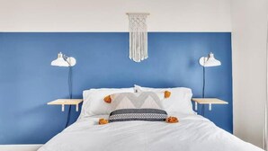 A bedroom with a blue accent wall, white bedding, two bedside tables with lamps, and a macrame hanging light fixture.