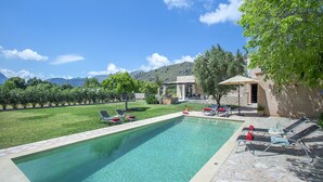 A rectangular swimming pool with clear water, surrounded by a stone deck. There are lounge chairs with red cushions and a table with chairs under a canopy. The pool area is adjacent to a grassy lawn with trees and a building with a stone facade. - VILLA VINYA FRARES - LUXURY RETREAT (Pollensa)