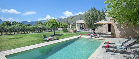 Ein rechteckiger Pool mit klarem Wasser, umgeben von einer Steindeck. Liegestühle mit roten Kissen und ein Tisch mit Stühlen unter einem Sonnensegel. Das Pool-Areal grenzt an eine Rasenfläche mit Bäumen und ein Gebäude mit einer Stein-Fassade.