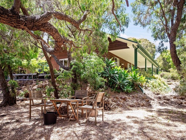 An outdoor seating area with wooden furniture and a table, surrounded by trees and plants.