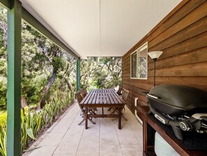 A covered patio with a wooden table and chairs, a grill, and a view of trees.