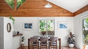 A dining area with a wooden ceiling, a brick wall, and a tiled floor. There are four chairs, a table, and two windows with views of greenery. - Ryma Cottage | Prevelly (Prevelly)