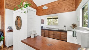 A kitchen with white brick walls, wooden ceiling beams, a wooden countertop, and a window with a view of trees. - Ryma Cottage | Prevelly (Prevelly)