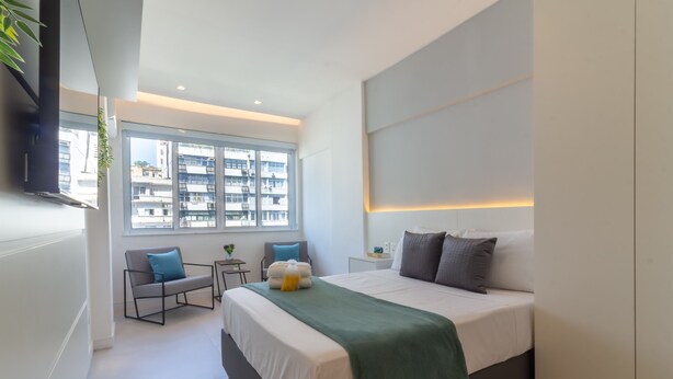 Une chambre moderne avec un grand lit, des tables de chevet, une fenêtre donnant sur la ville et un téléviseur à écran plat fixé au mur.