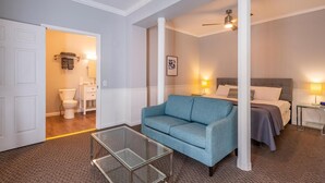 A hotel room with a blue sofa, a glass coffee table, a bed with a grey headboard, and a bathroom visible through an open door. - Luxury Hot tub Suite w/ In-Room Hot Tub & King Bed 1BR, 1BA (Nashville)