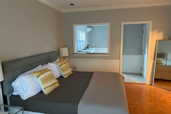 King bed with gray headboard, yellow patterned pillows, and a view into the white kitchen through an interior window.