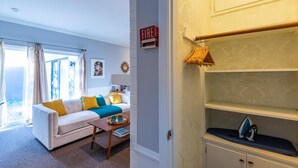 A living room with a white sofa, a coffee table, and a bookshelf. There is a fire alarm on the wall and a window with curtains. - Luxury Hot tub Suite w/ In-Room Hot Tub & King Bed 1BR, 1BA (Nashville)