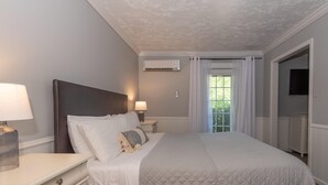 A bedroom with a large bed, two bedside tables with lamps, an air conditioning unit, a window with curtains, and a television on the wall. - Luxury Hot tub Suite w/ In-Room Hot Tub & King Bed 1BR, 1BA (Nashville)