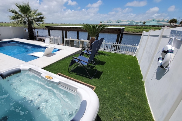Hot tub bubbles beside a rectangular concrete pool, blue reclining chairs on artificial turf near waterway dock.