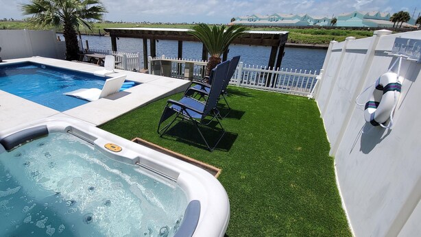 A swimming pool, hot tub, and lounge chairs on a grassy deck with a view of a body of water and a dock. - Island Waterfront 3/2 Private Pool & Hot Tub with Boat Dock (Corpus Christi)