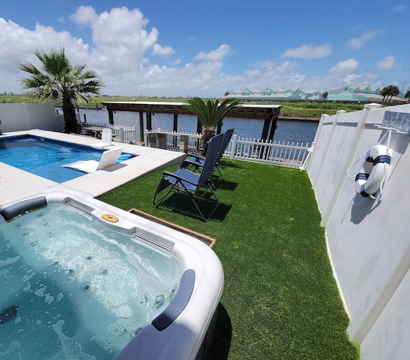 Una piscina, un jacuzzi y sillas de descanso en una terraza con césped, con vistas a un cuerpo de agua y un muelle.