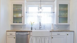 A kitchen with white cabinets, a stainless steel dishwasher, a granite countertop, a window with sheer curtains, and a chandelier. - Walkable SoCo Bungalow (Austin)