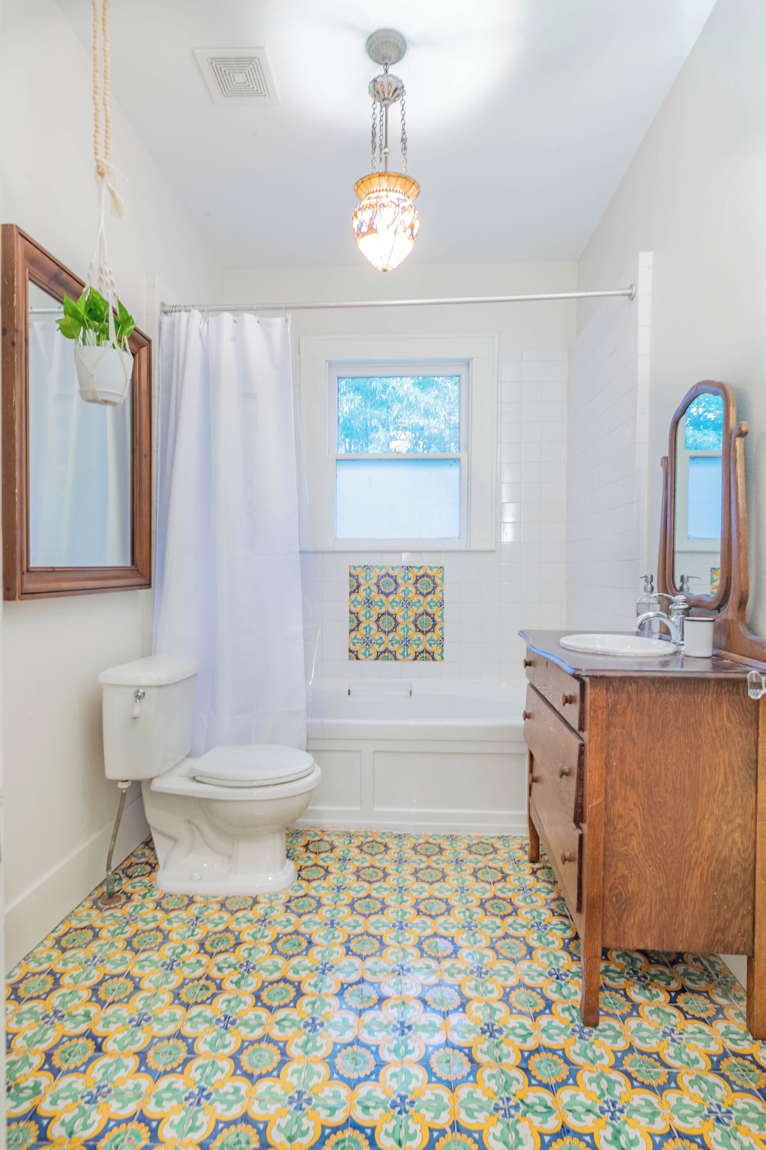 Une salle de bain équipée de toilettes, d’une baignoire et d’un lavabo. Il y a une fenêtre au-dessus de la baignoire et un lustre suspendu au plafond.