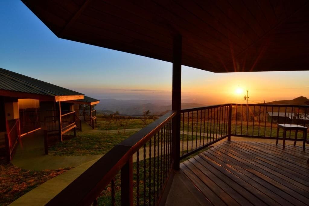 A wooden deck with a metal railing overlooking a landscape with a sunset.
