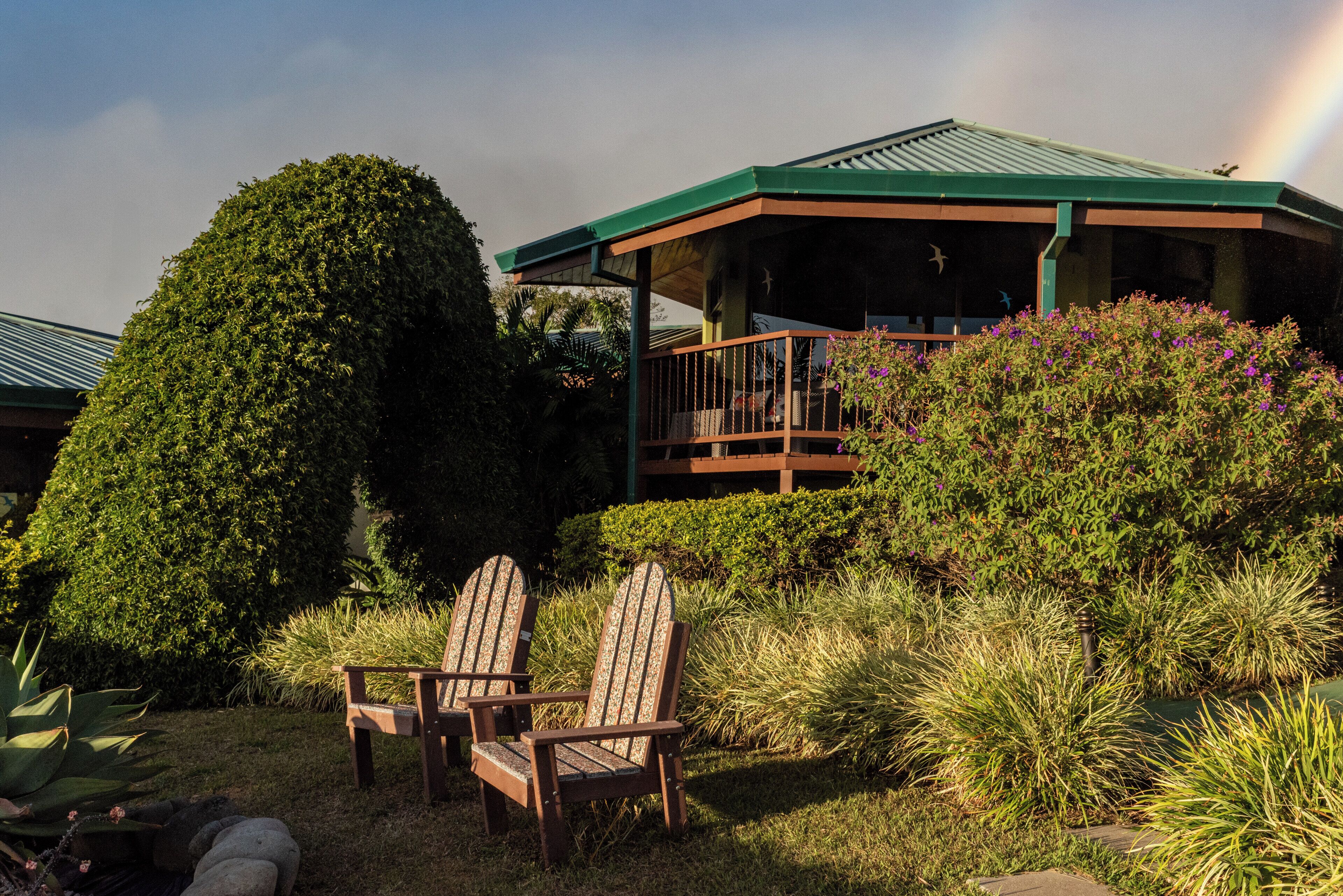 A gazebo with a green roof, surrounded by lush greenery and two Adirondack chairs.