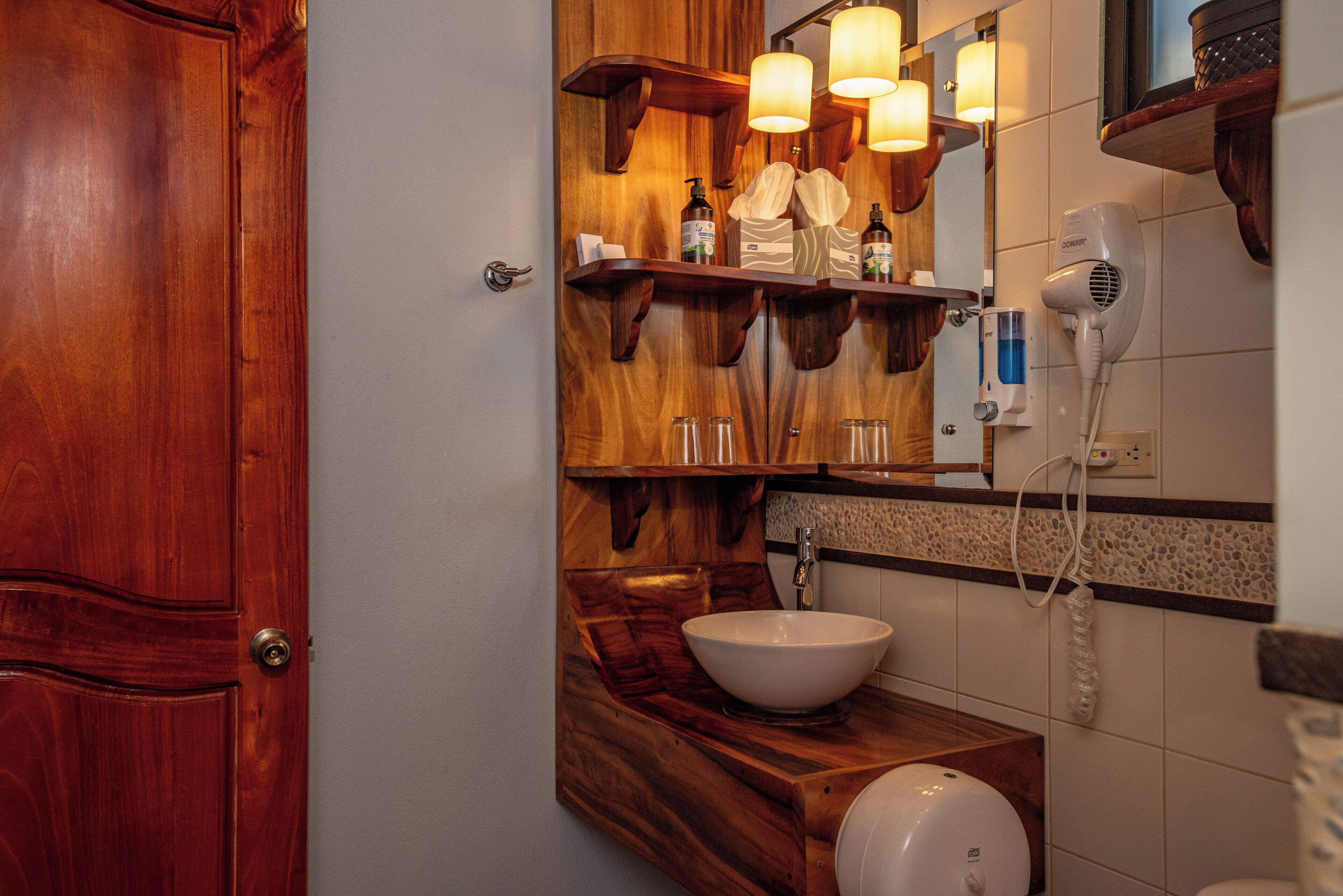 Ein Badezimmer mit einem Holzbadezimmer-Möbelstück, einem weißen Waschbecken, einem Spiegel mit Holzregalen und einem Haartrockner an der Wand.