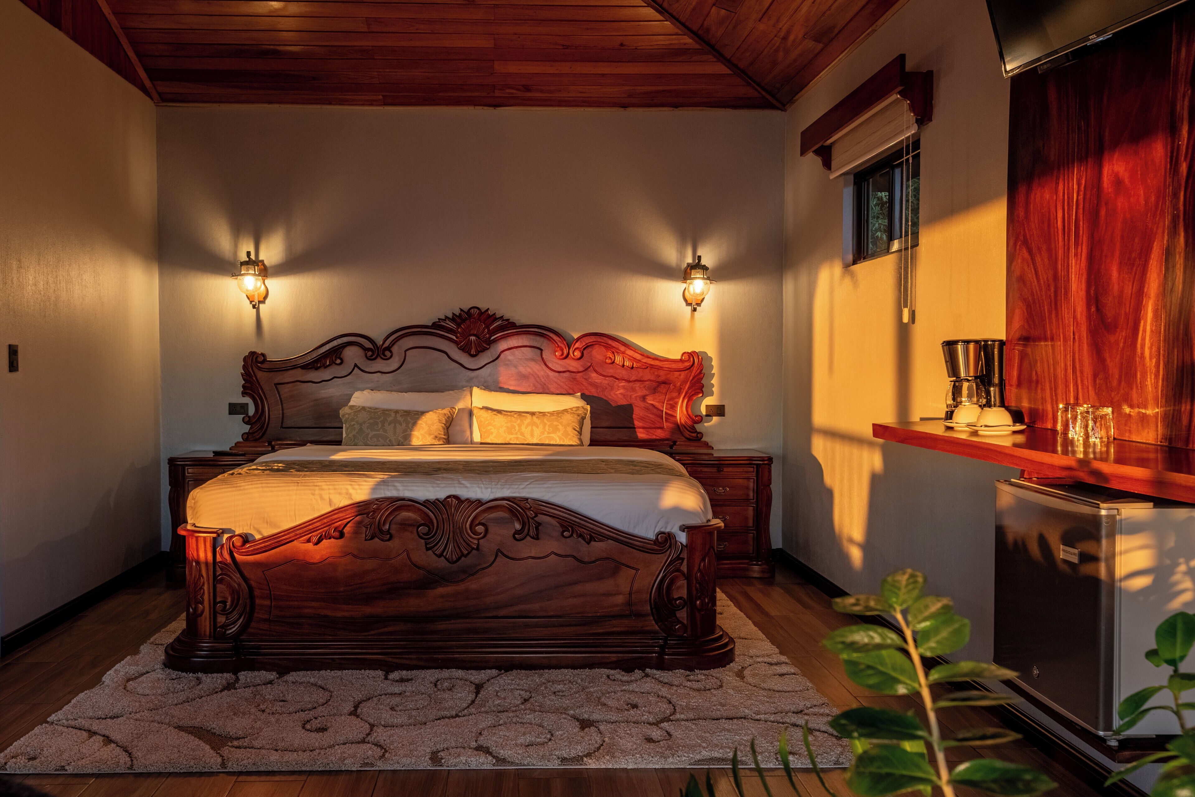 A wooden bed with a headboard, two wall sconces, a bedside table, and a small refrigerator.