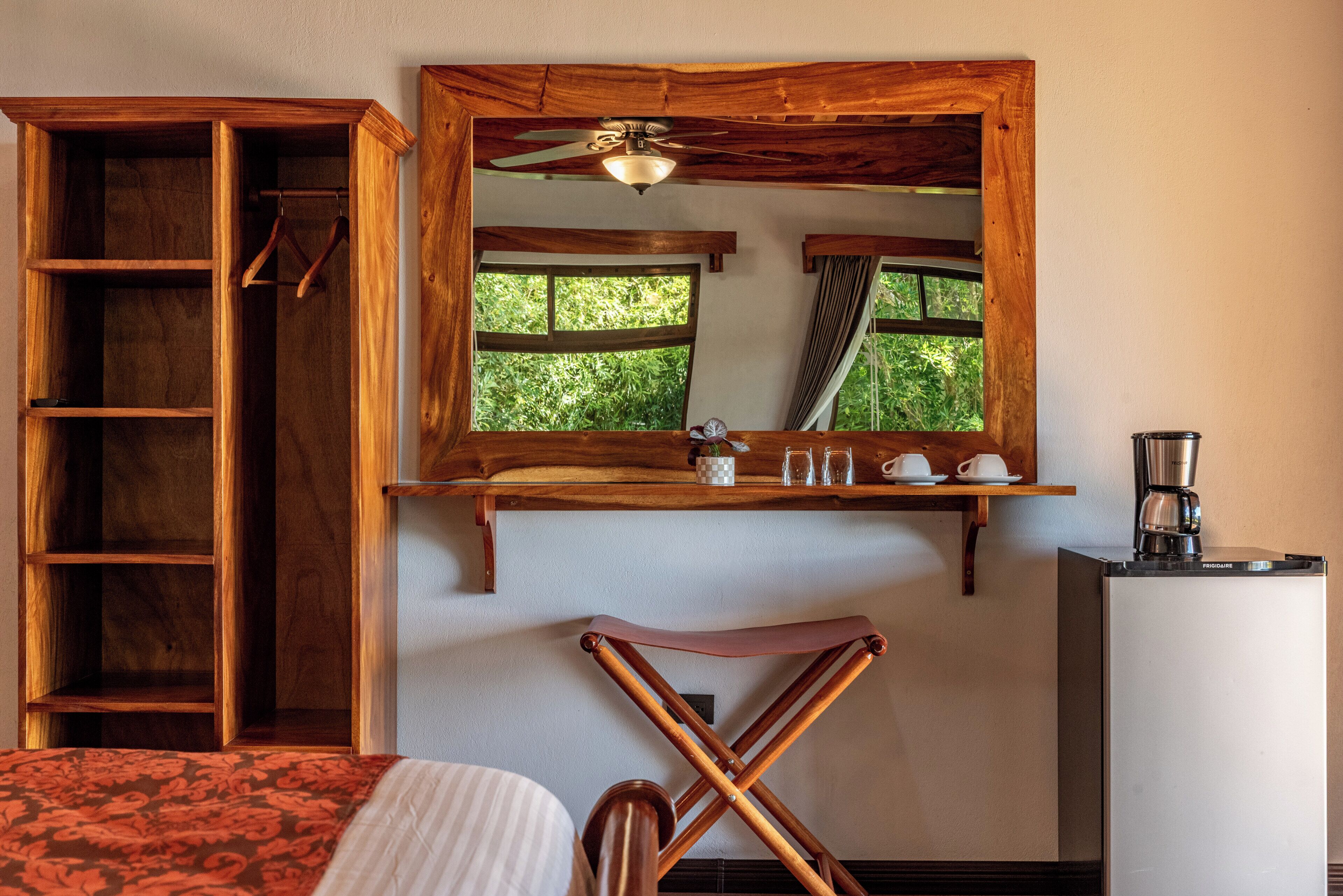 A room with a bed, a wooden wardrobe, a mirror, a ceiling fan, a window, and a coffee maker.