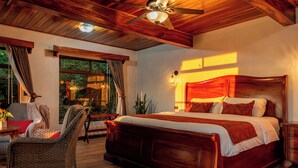 A bedroom with a wooden ceiling, a large bed, a wicker chair, and a view of greenery outside. - Hotel Trópico Monteverde (Monteverde)