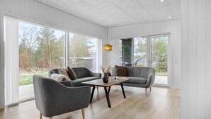 Amplia sala de estar con dos sofás grises, una mesa de centro de madera y una lámpara colgante. Grandes ventanas ofrecen vistas al exterior.