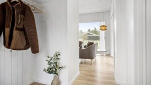 Un pasillo con un perchero, un florero de mimbre con plantas verdes y vistas a una habitación con una silla gris y una lámpara colgante.