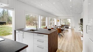 Una cocina moderna con una isla grande, armarios blancos y vistas al jardín trasero a través de ventanas amplias.