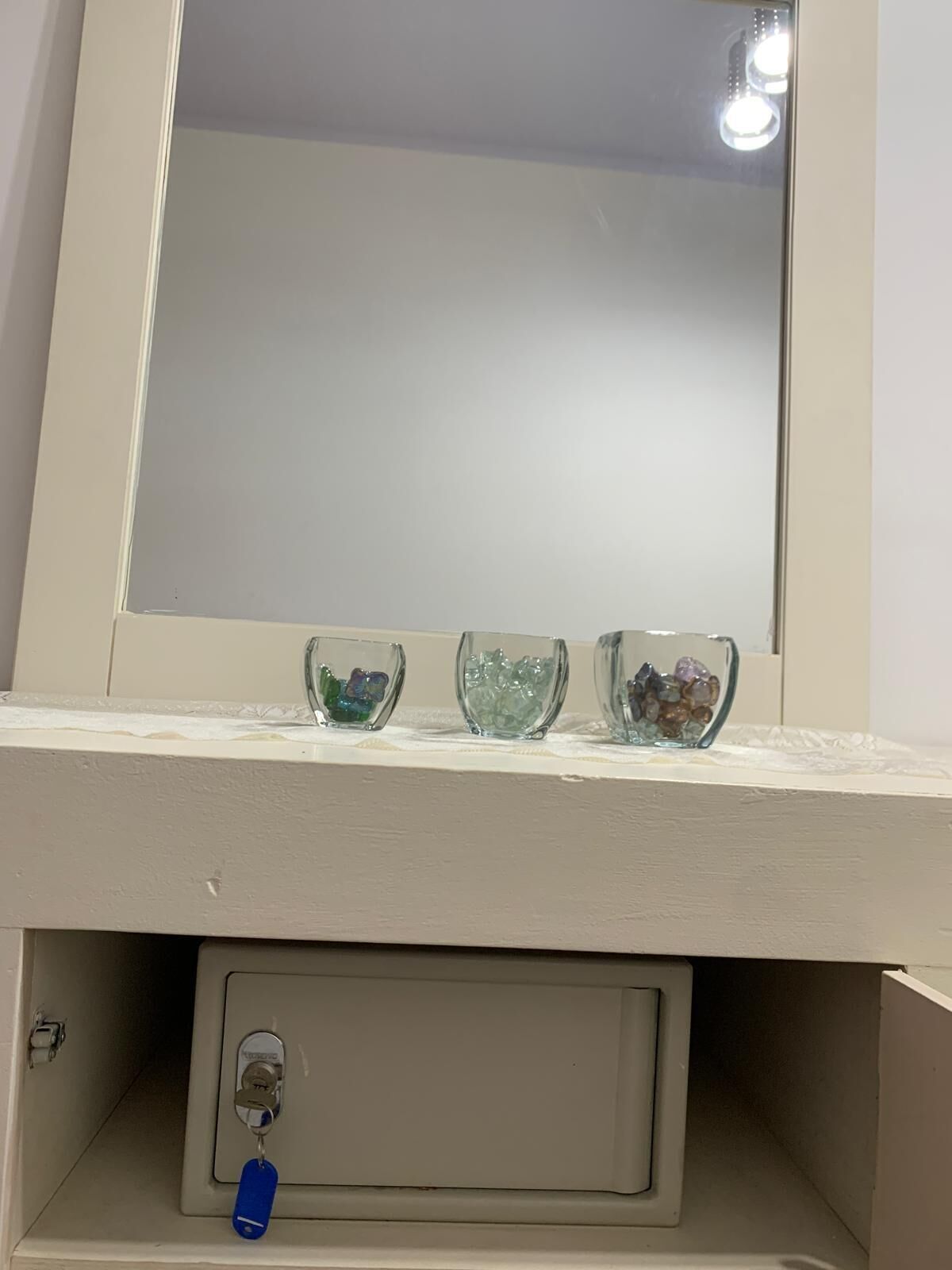 Un tocador de baño con espejo, tres recipientes de vidrio con piedras decorativas y un mueble pequeño con cerradura.