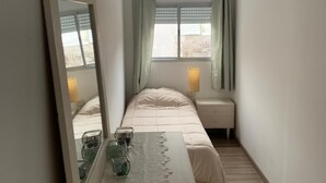 Una habitación estrecha con una cama, una mesita de noche con lámpara, un espejo y una ventana con cortinas.