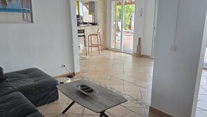 Un vaste espace de vie comprenant un canapé noir, une petite table en bois et un coin cuisine visible à travers une porte ouverte.