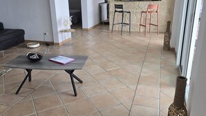 Un vaste espace de vie doté d’un sol carrelé, d’un canapé noir, d’une petite table sur laquelle se trouvent un bol et un livre, ainsi que d’un coin cuisine avec un comptoir et deux chaises.