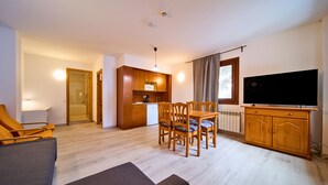 Een moderne woonkamer met een bank, een eettafel en stoelen, een keuken met een magnetron en kastjes, en een flatscreen televisie aan de muur.