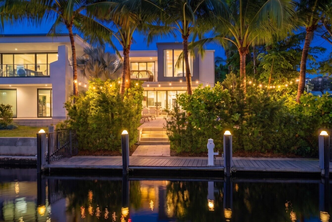 A modern two-story house with a pool, surrounded by lush greenery and palm trees.