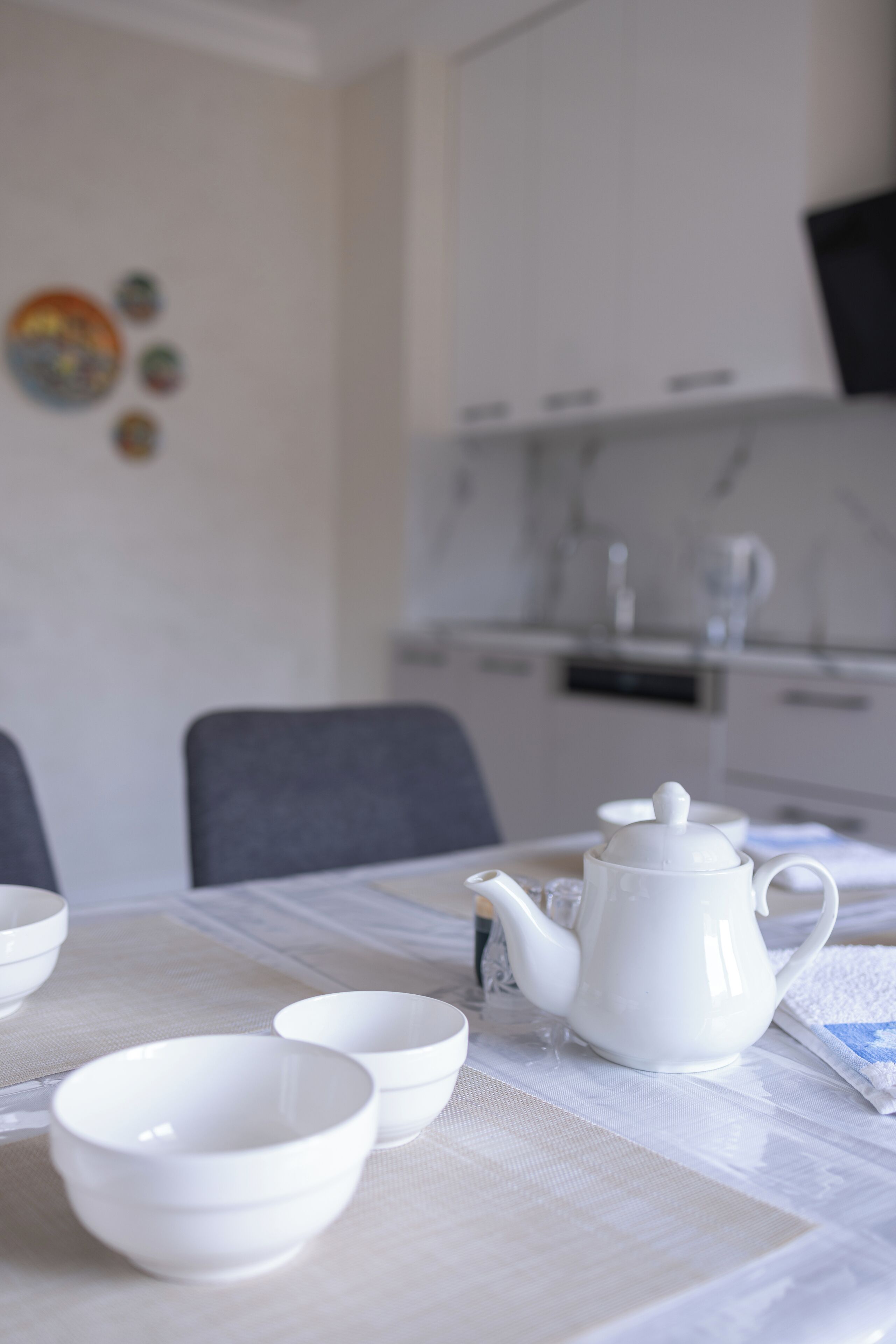 A dining table with white ceramic tea set and bowls, a white tablecloth, and a kitchen in the background.